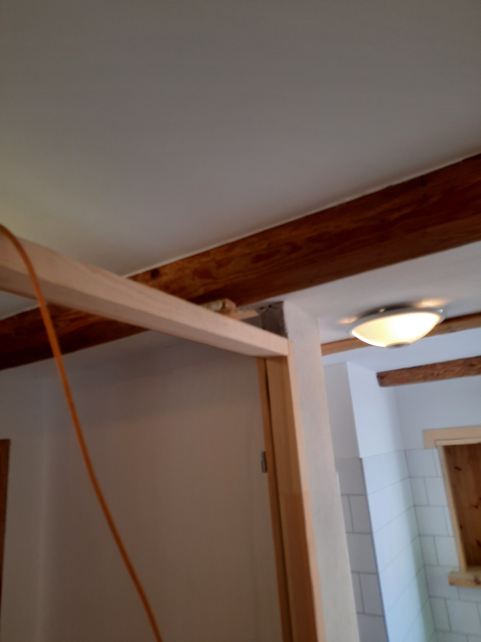 Wnętrze w trakcie przebudowy, widoczna drewniana konstrukcja ścianki działowej, fragment instalacji elektrycznej w postaci pomarańczowego kabla oraz wykończona łazienka z białymi płytkami...