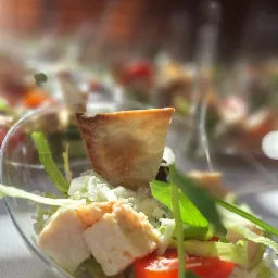 Delikatna sałatka w przezroczystej miseczce z kawałkami kurczaka, pomidorem, sałatą i grzanką, oświetlona naturalnym światłem, z rozmytym tłem zastawionego stołu.