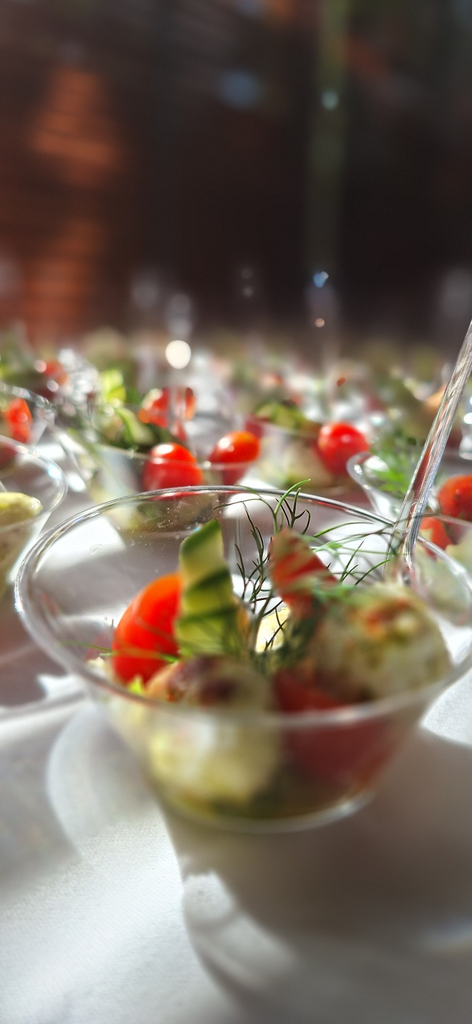 Mini porcje sałatek z pomidorami, ogórkami, serem i ziołami w przezroczystych naczyniach, ustawione na białym obrusie, oświetlone naturalnym światłem.