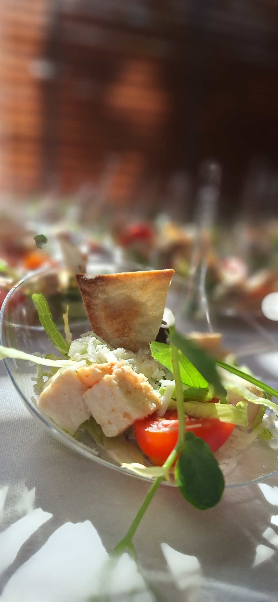 Delikatna sałatka w przezroczystej miseczce z kawałkami kurczaka, pomidorem, sałatą i grzanką, oświetlona naturalnym światłem, z rozmytym tłem zastawionego stołu.