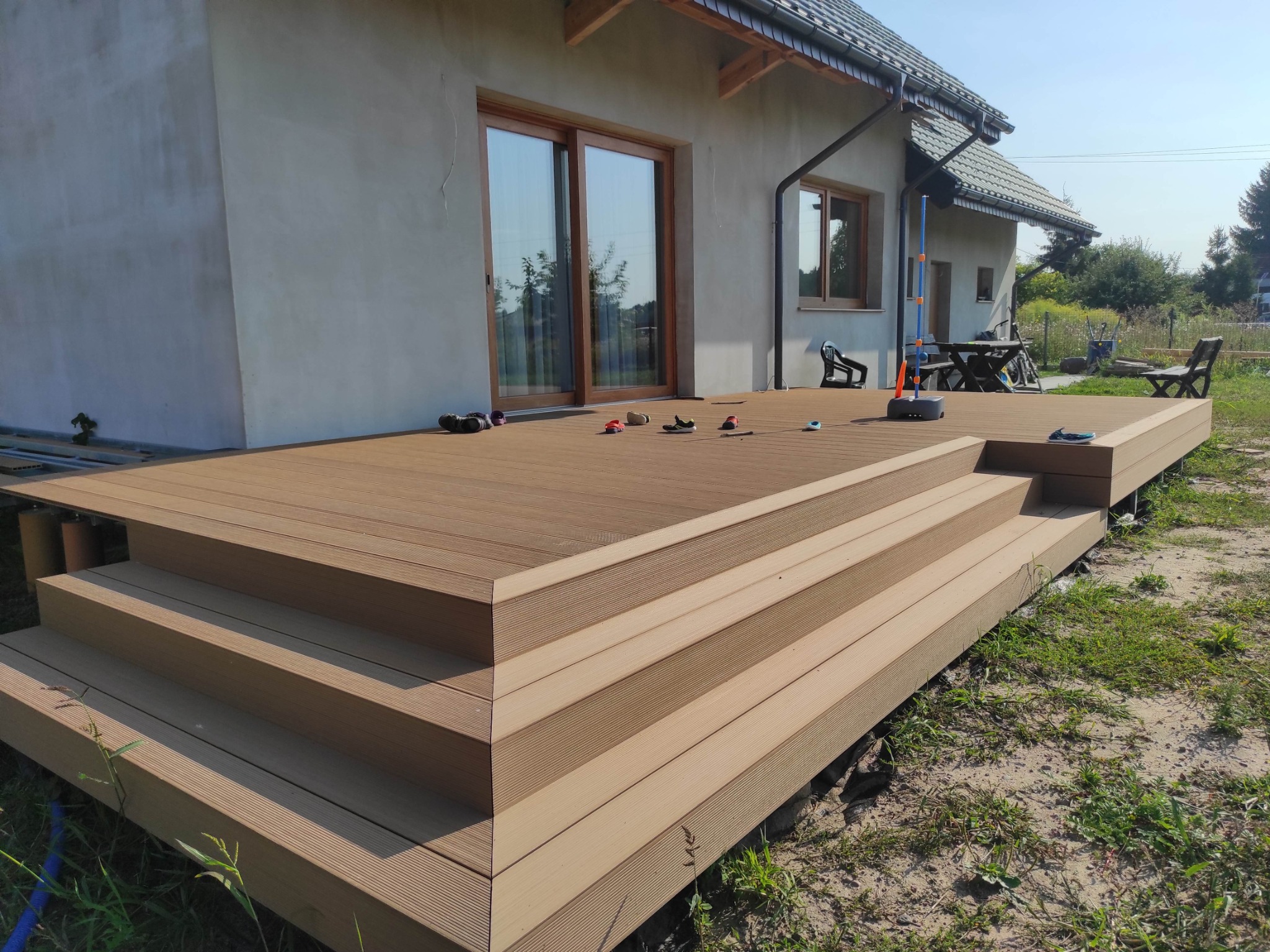 Nowoczesny taras z desek kompozytowych z trzema schodkami prowadzącymi do jasnego domu z dużymi przesuwnymi drzwiami tarasowymi, otoczony zielenią i pozostawionymi butami.