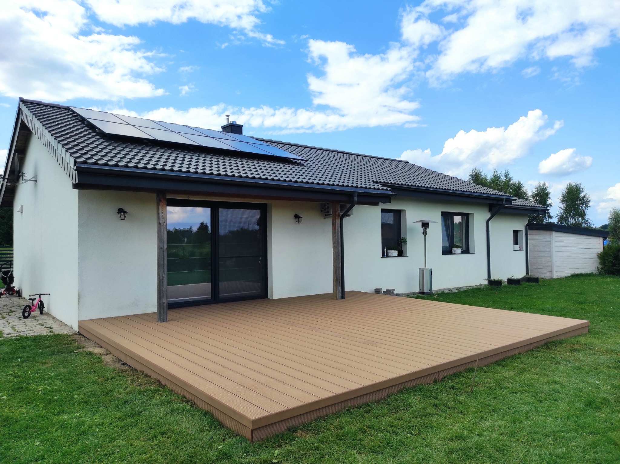 Gotowy taras z desek kompozytowych przy białym domu z panelami słonecznymi w Nikielkowie, widok z trawnika, błękitne niebo z chmurami.