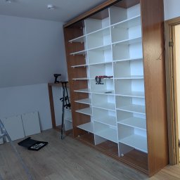 Przedsiębiorstwo Usługowe MAX Wojciech Daniluk - Montaż regału z półkami w Tczewie. Widoczna drabina, narzędzia i elementy mocujące. Regał w trakcie składania, z widocznymi klamrami montażowymi.