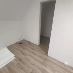 Jasne panele podłogowe ułożone w pokoju ze skośnym sufitem, widoczne kable przy ścianie i białe podkłady pod panele. Widok na sąsiednie pomieszczenie.