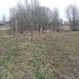 Montaż słupków drewnianych do ogrodzenia na zewnątrz, widoczne druty, w tle krzewy i zabudowania.