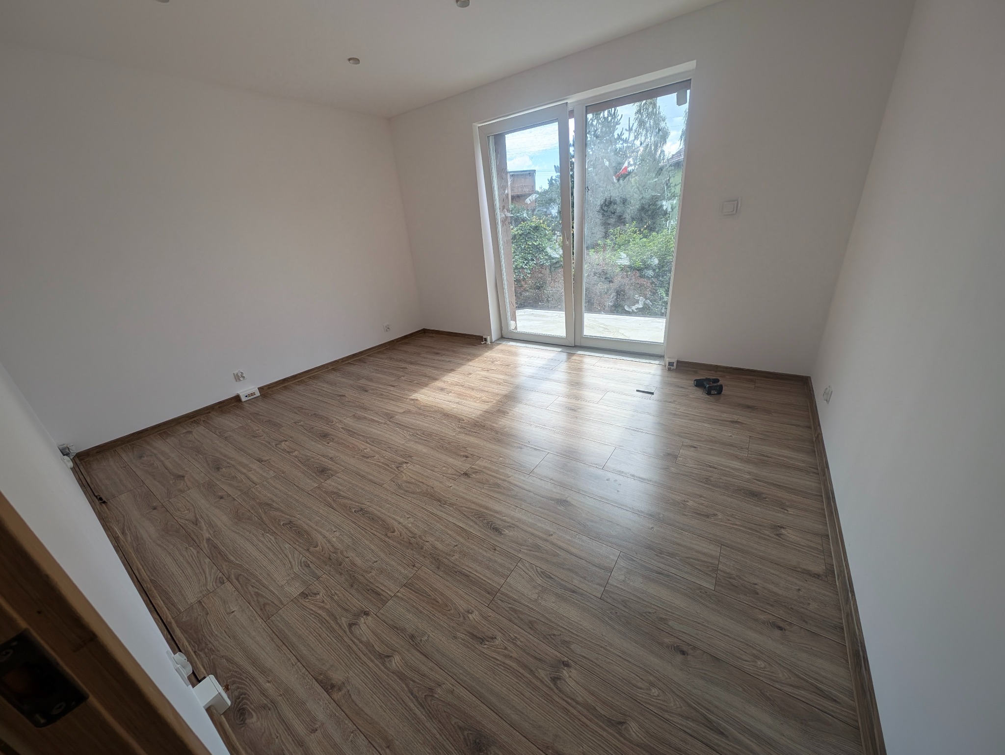 Jasne panele podłogowe w nowym pokoju z dużym oknem balkonowym. Widoczne narzędzia przy ścianie. Naturalne światło podkreśla wzór drewna.