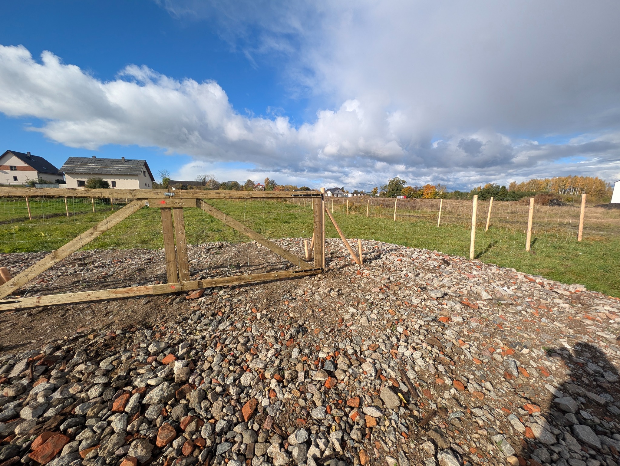 W trakcie budowy ogrodzenia z siatki: drewniana brama wejściowa z siatką i słupki w ziemi na tle domów i błękitnego nieba z chmurami. Widok z bliska na gruz.