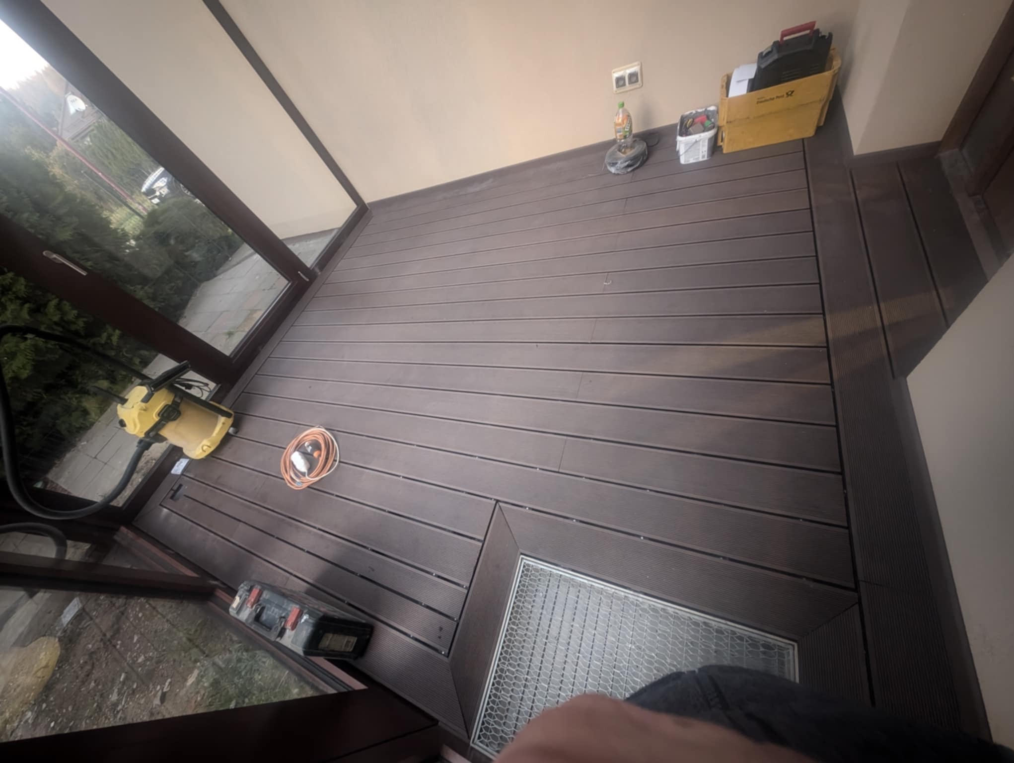 Ciemnobrązowy taras kompozytowy z widocznym odpływem liniowym i narzędziami pracy, częściowo zadaszony, z widokiem na zieleń przez okno.