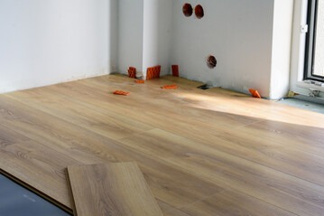 Układanie paneli podłogowych w rogu pomieszczenia. Widoczne pomarańczowe elementy instalacji elektrycznej na ścianie i fragmenty paneli na podłodze. Jasne wnętrze w trakcie prac.