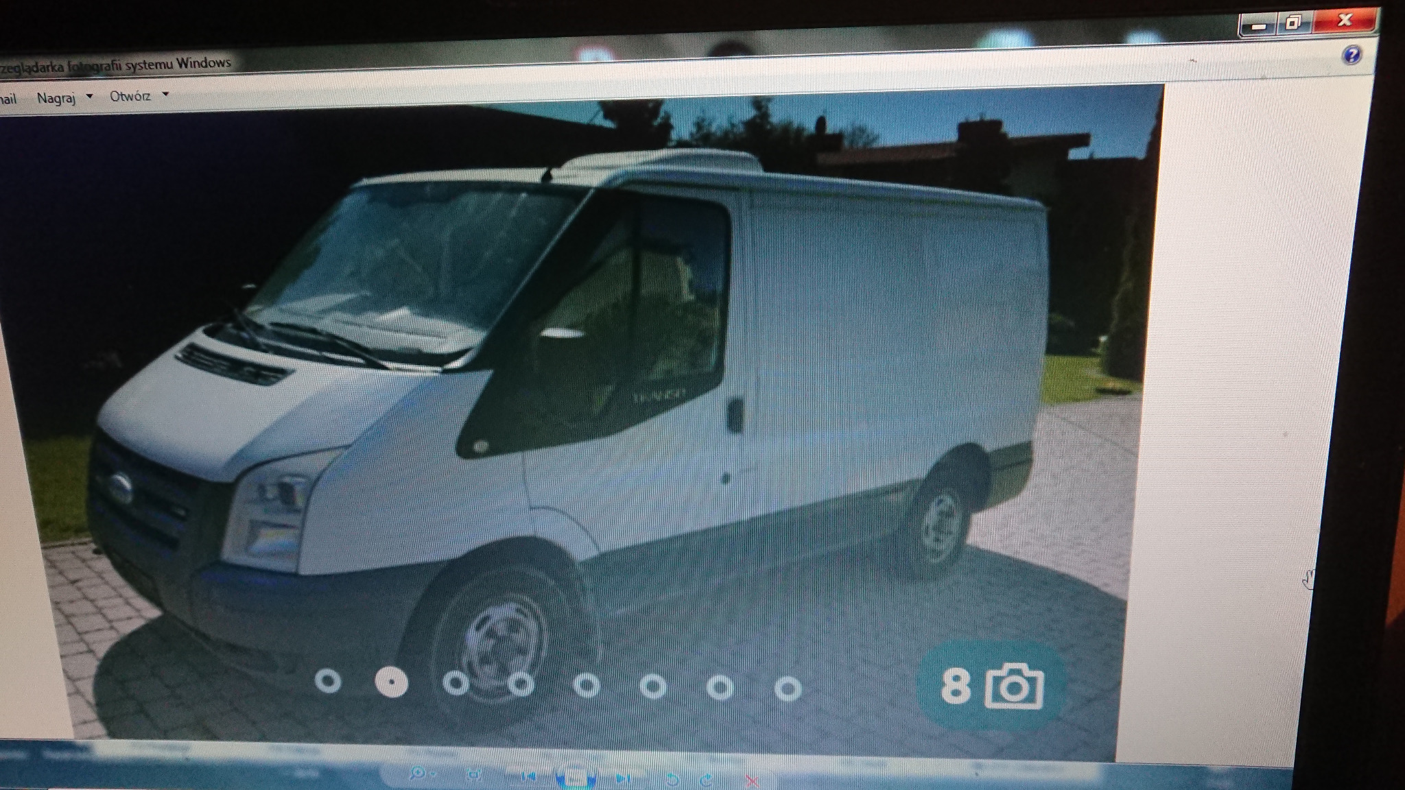 Biały furgon Ford Transit z nadbudówką dachową, zaparkowany na brukowanej powierzchni przed budynkiem z zielonym trawnikiem w tle.