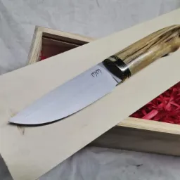 Ręcznie wykonany nóż z jasnym drewnianym uchwytem, zdobionym metalowymi pierścieniami, leży na drewnianej desce w otwartym drewnianym pudełku wypełnionym czerwoną bibułą.