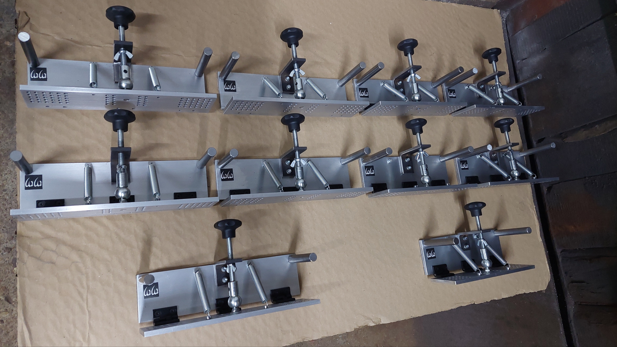Szereg srebrnych, metalowych uchwytów z regulacją wysokości, sprężynami i otworami, ułożonych na brązowym papierze, widoczne logo firmy.