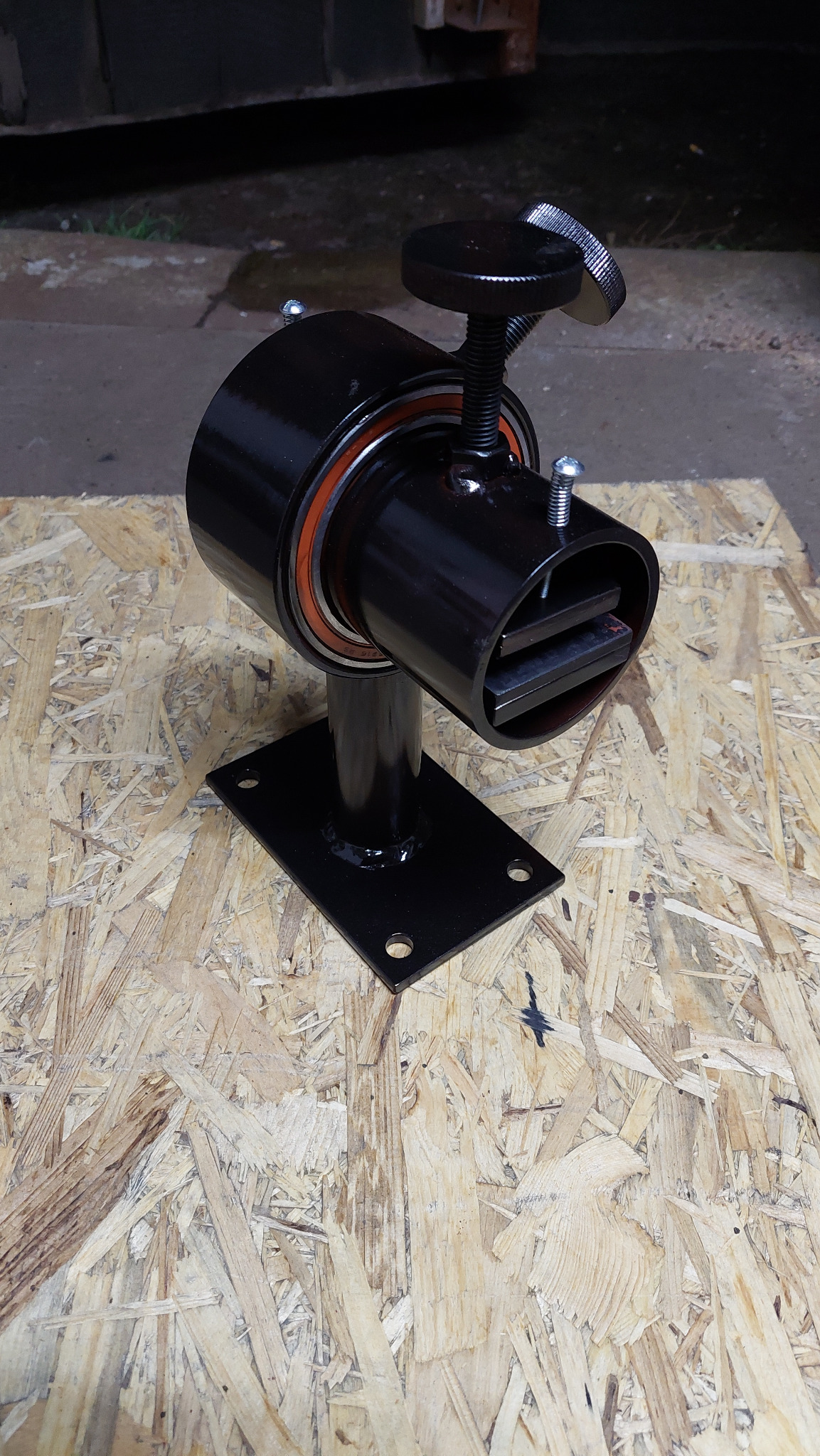 Czarny, metalowy uchwyt z regulacją śrubową, zamontowany na płycie OSB, widoczne elementy łożyska i dwa metalowe pręty wewnątrz uchwytu.