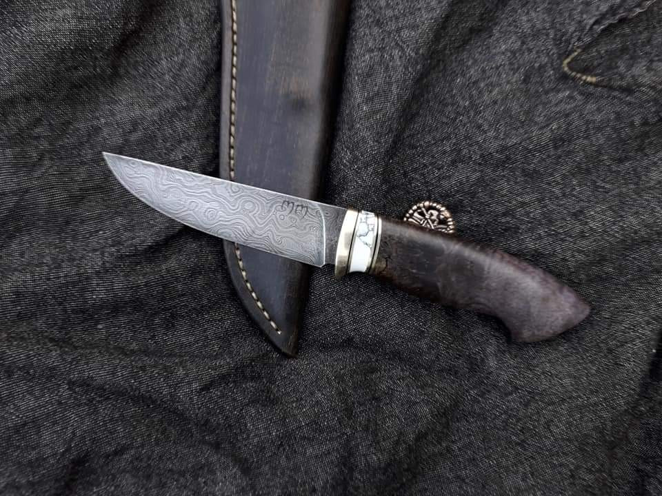 Ręcznie wykonany nóż z damasceńskiej stali, z drewnianą rączką i białymi wstawkami, leżący obok skórzanej pochwy na tle ciemnego materiału.