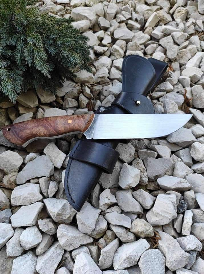 Nóż myśliwski ze stalowym ostrzem i drewnianą rękojeścią w skórzanej pochwie, ułożony na tle z białych kamieni i gałązki iglaka.