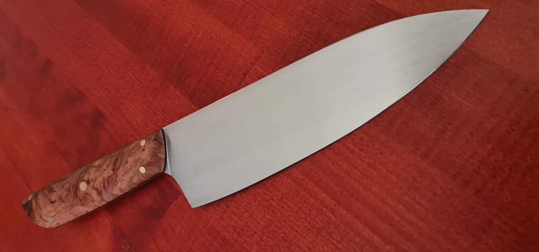 Nóż szefa kuchni z ostrzem ze stali nierdzewnej i rzeźbioną rękojeścią z jasnego drewna na czerwonym drewnianym blacie.