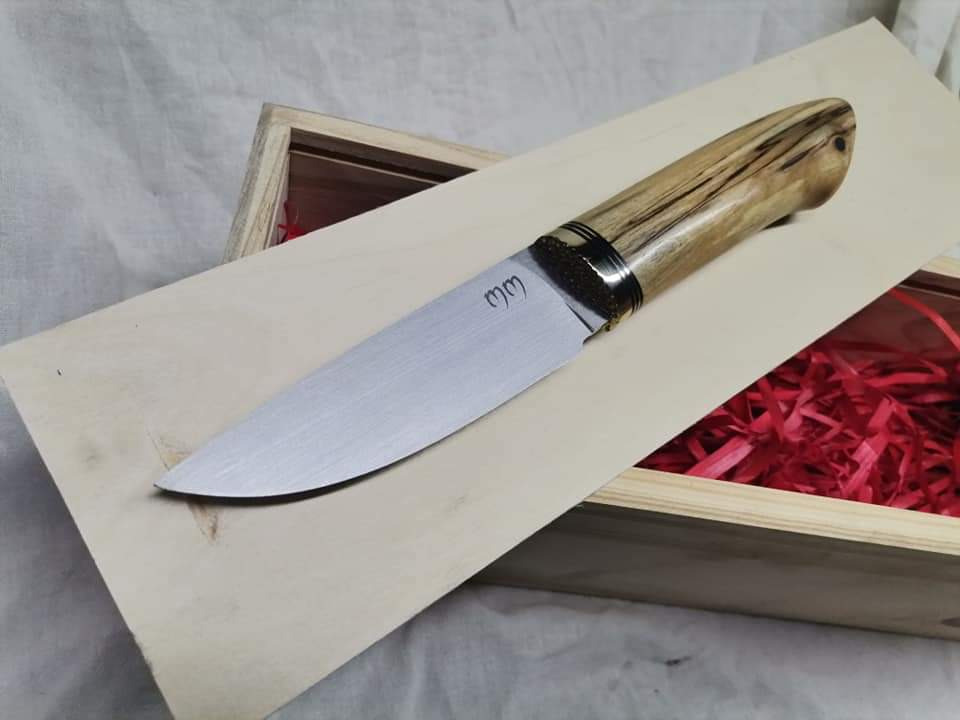 Ręcznie wykonany nóż z jasnym drewnianym uchwytem, zdobionym metalowymi pierścieniami, leży na drewnianej desce w otwartym drewnianym pudełku wypełnionym czerwoną bibułą.