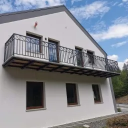 Nowoczesny dom z balkonem z czarną, metalową balustradą o prostym wzorze, widok z zewnątrz w słoneczny dzień.