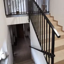 Nowoczesne schody z czarną metalową balustradą, widok z góry na dolny bieg schodów i antresolę z rośliną doniczkową.