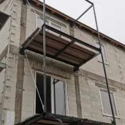 Konstrukcja metalowa balkonu z drewnianym wypełnieniem, przymocowana do surowej ściany budynku z widocznymi oknami, zabezpieczona folią budowlaną.