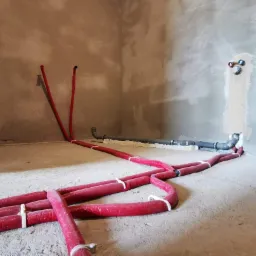 Surowe wnętrze z instalacją hydrauliczną: czerwone rury grzewcze na podłodze, szare rury kanalizacyjne przy ścianie, ściany w stanie deweloperskim.