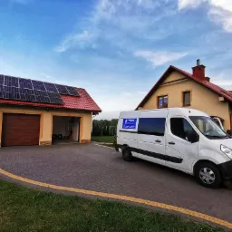 Biały van serwisowy zaparkowany przed garażem z panelami słonecznymi na dachu, w tle dom jednorodzinny z czerwonym dachem.