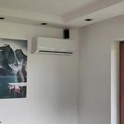 Nowoczesny klimatyzator ścienny zamontowany w jasnym salonie z obrazami przedstawiającymi naturę, widoczne również okno i roślina doniczkowa.