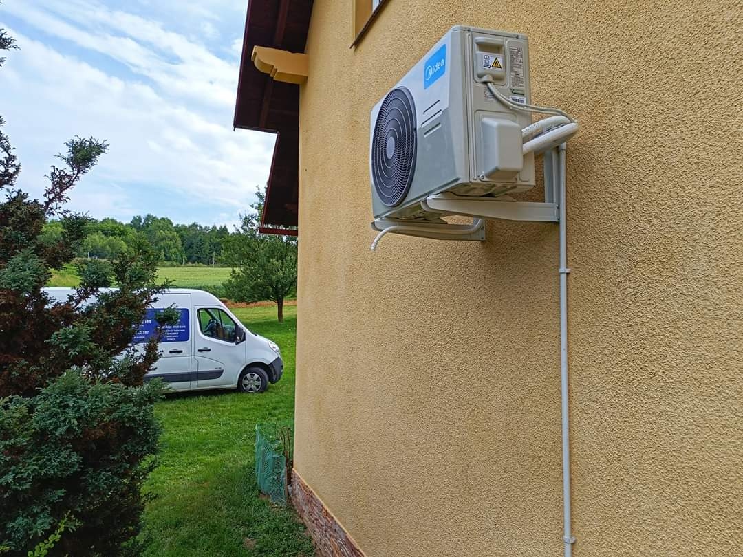 Zewnętrzna jednostka klimatyzacji Midea zamontowana na żółtej elewacji domu, z widoczną instalacją i samochodem serwisowym w tle.