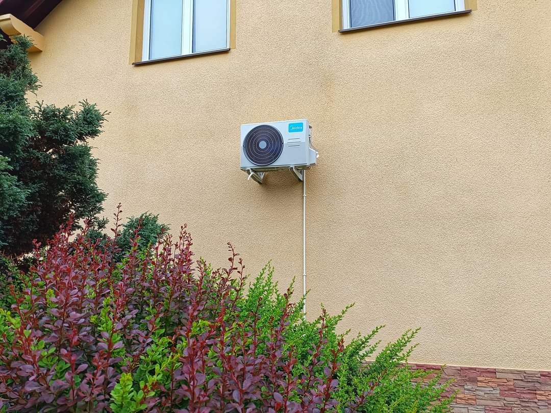 Klimatyzator Midea zamontowany na elewacji budynku z oknami, widoczny fragment instalacji i ozdobny krzew na pierwszym planie.