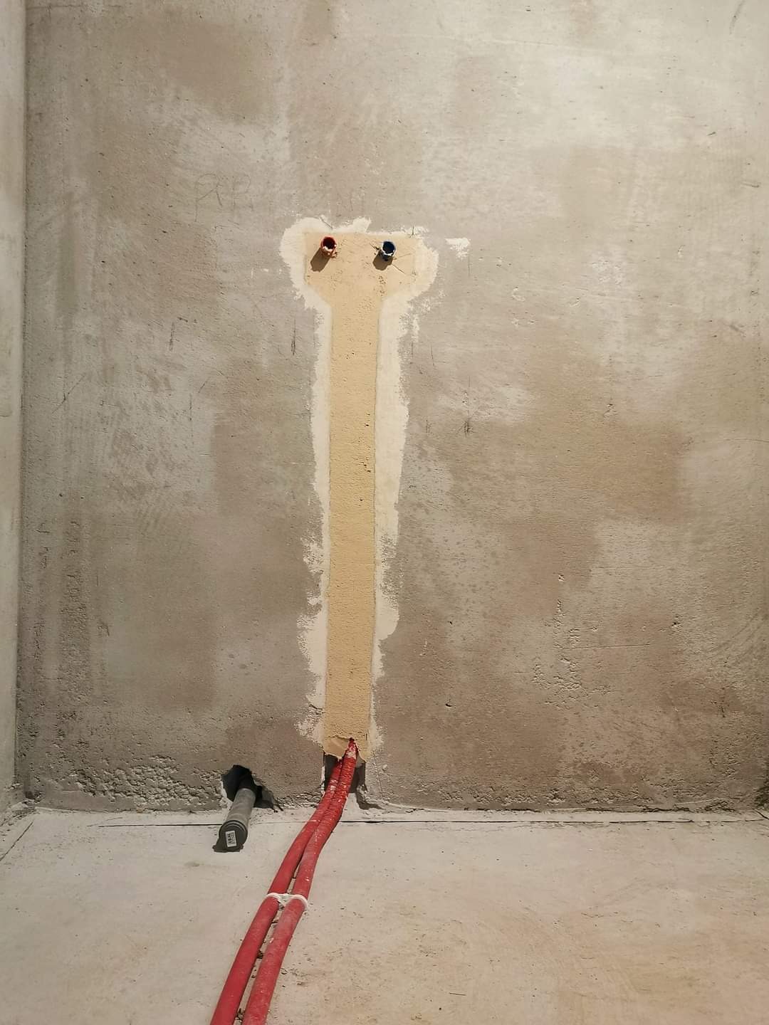 Surowa ściana z wyżłobionym pionowym kanałem wypełnionym zaprawą, z wystającymi rurami PEX w kolorze czerwonym i otworami na instalację wodną z zaślepkami w kolorze czerwonym i niebieskim.