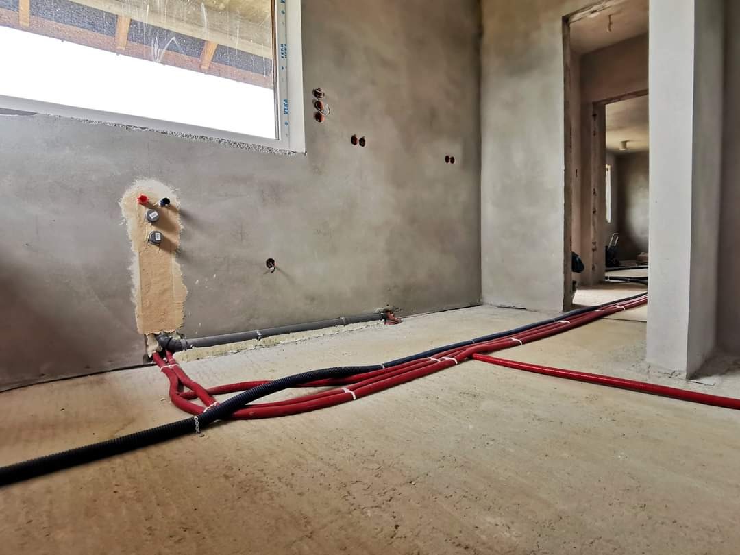 Surowy stan wnętrza z widoczną instalacją wodną i elektryczną: czerwone i czarne rury prowadzone po podłodze, wyprowadzenia instalacji wodnej na ścianie pod oknem, puszki elektryczne w ścianie.