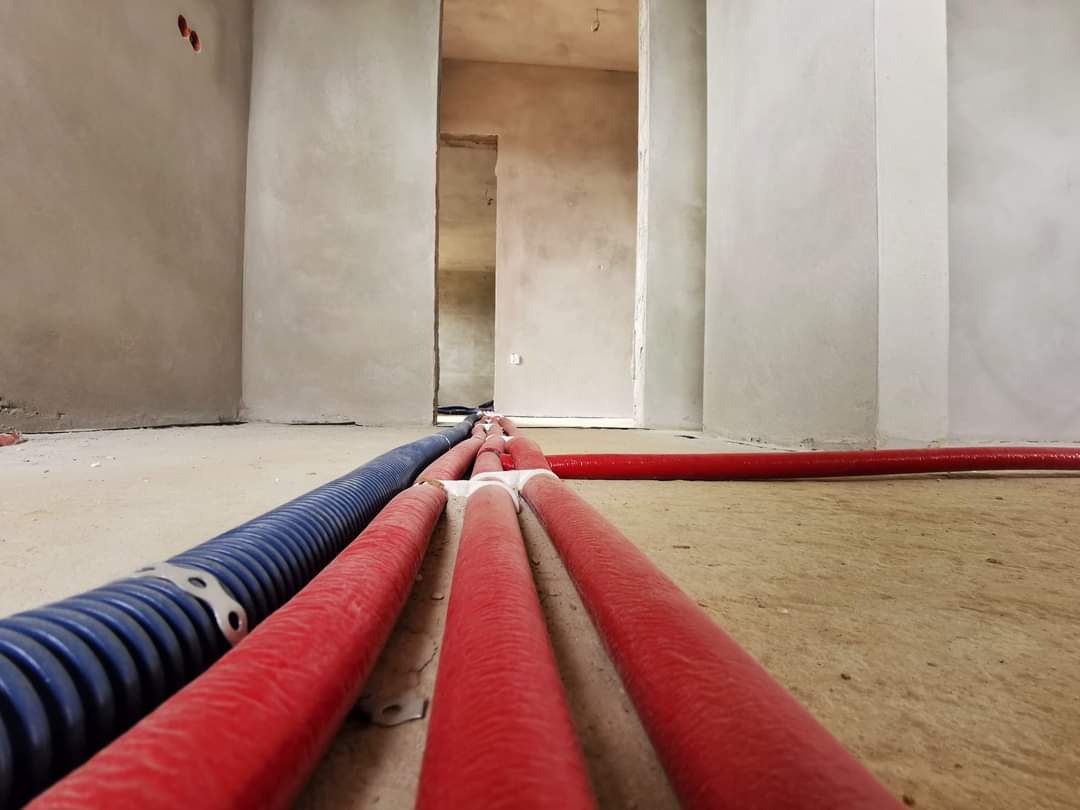 Niebieska karbowana i czerwone gładkie rury prowadzone po surowej posadzce w trakcie prac instalacyjnych wewnątrz pomieszczenia w stanie deweloperskim.