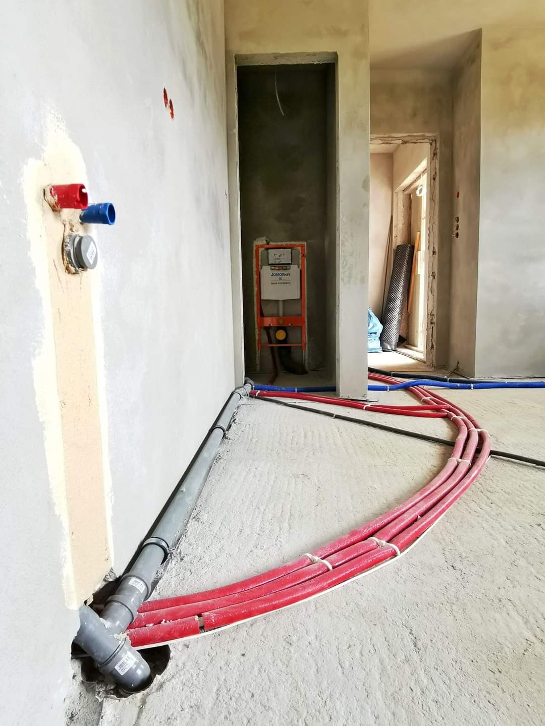 Instalacja wodna i kanalizacyjna w surowym stanie: czerwone rury ogrzewania podłogowego, szara rura kanalizacyjna i niebieskie rury wodne w nowo budowanym pomieszczeniu z widoczną instalacją...