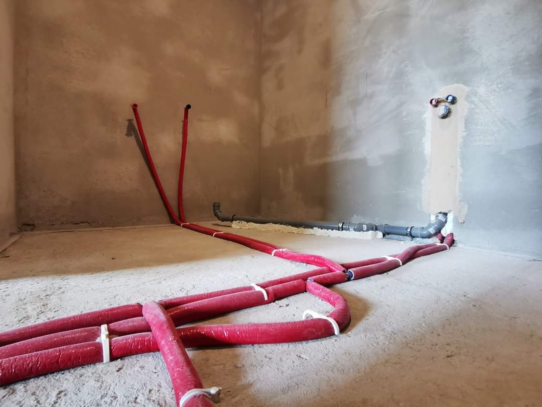 Surowe wnętrze z instalacją hydrauliczną: czerwone rury grzewcze na podłodze, szare rury kanalizacyjne przy ścianie, ściany w stanie deweloperskim.