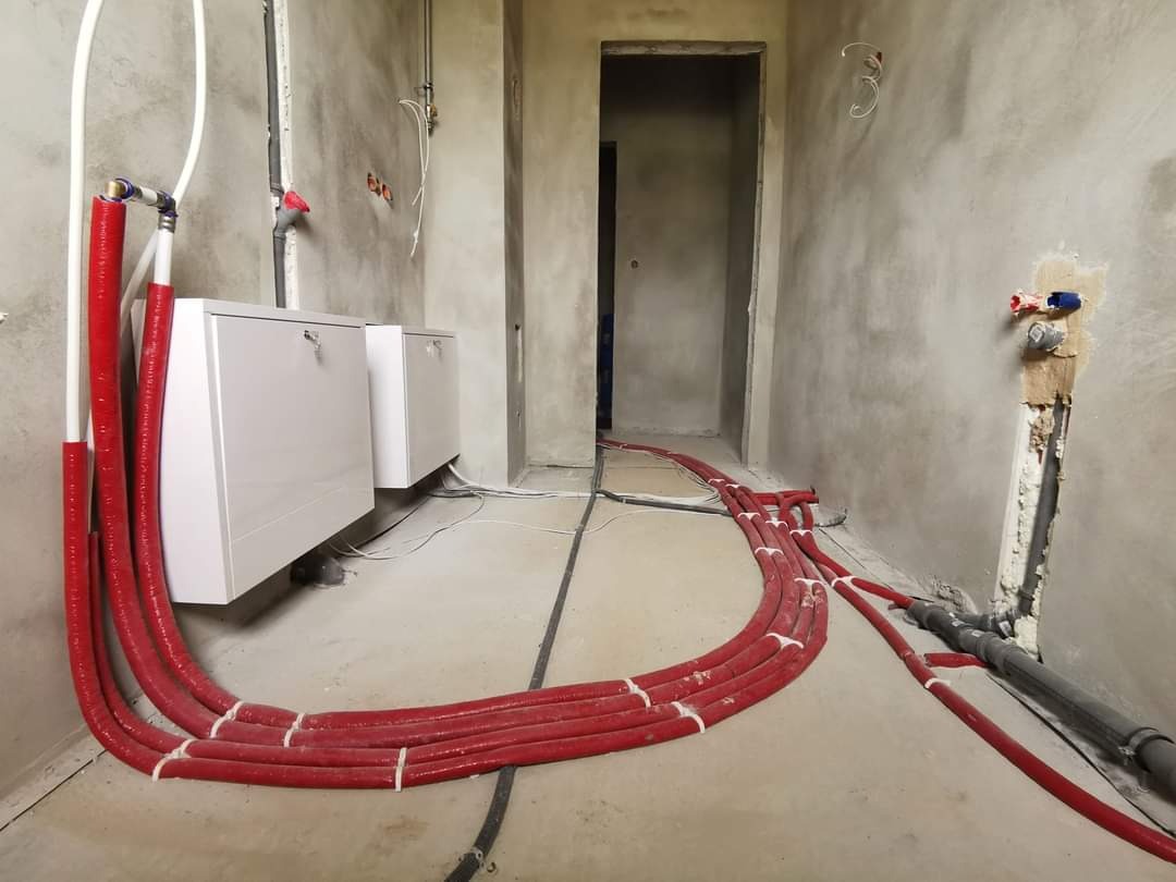 Surowe wnętrze w trakcie instalacji ogrzewania podłogowego: czerwone rury grzewcze rozłożone na posadzce, białe szafki rozdzielcze na ścianie, widoczne przyłącza wodne i kanalizacyjne.