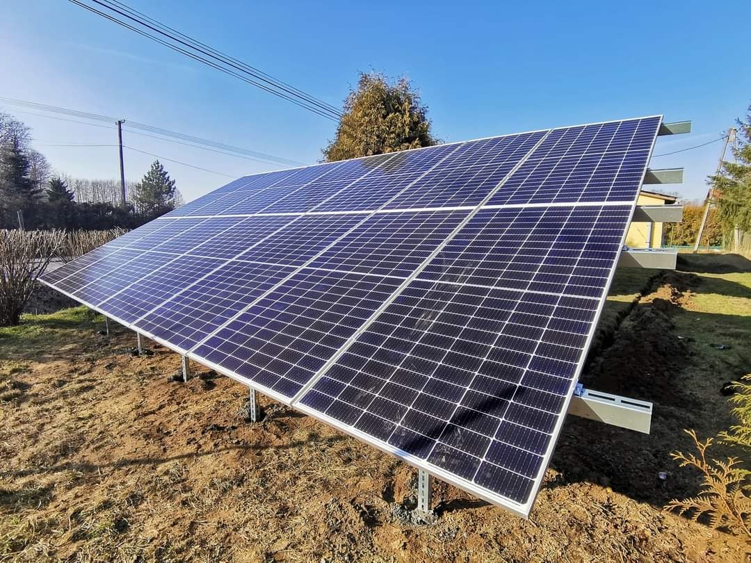 Panel słoneczny na metalowej konstrukcji, świeżo zamontowany na ziemi z widocznym budynkiem w tle, słoneczny dzień.