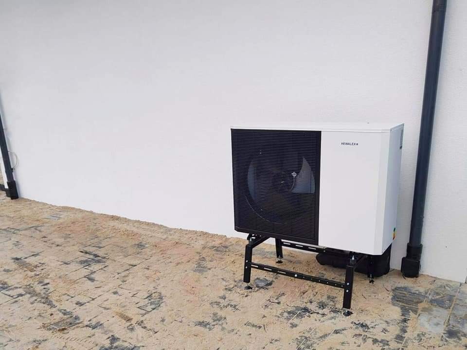 Biała pompa ciepła Hewalex na czarnym stelażu, zainstalowana przy białej ścianie, widoczne rury odprowadzające wodę i brudna podłoga.