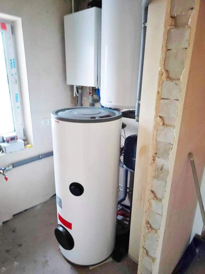 Bojler elektryczny z dwoma otworami inspekcyjnymi i czerwonym paskiem, zamontowany w surowym pomieszczeniu z widoczną instalacją grzewczą i oknem.