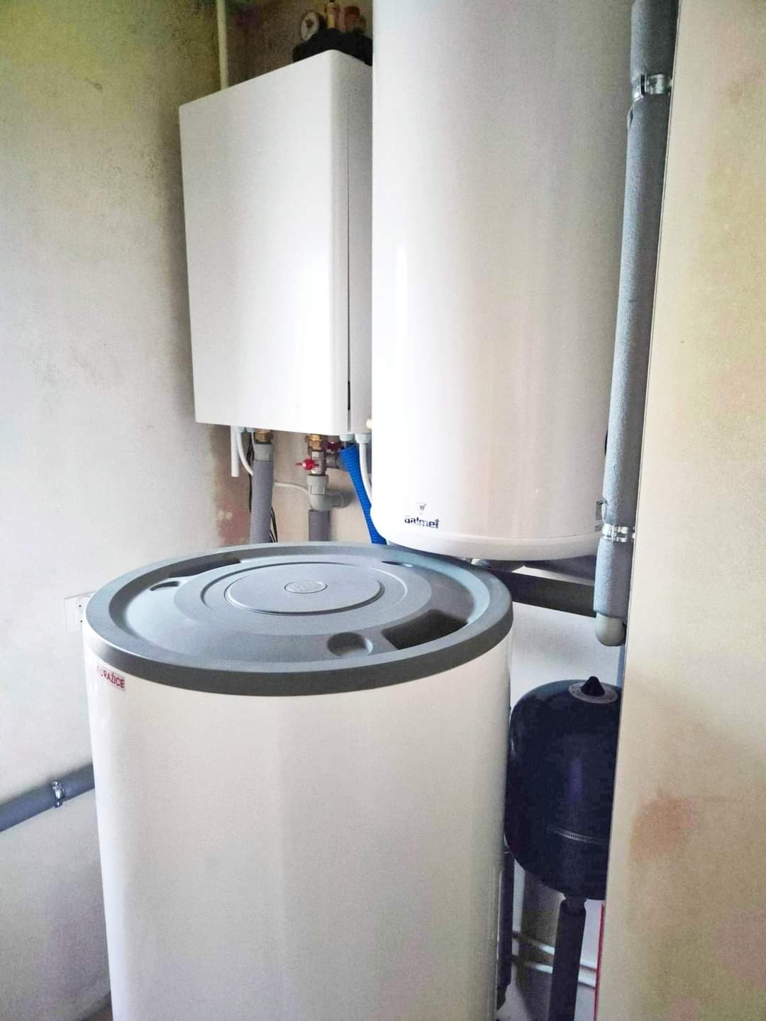 Kompaktowa kotłownia z białym zasobnikiem ciepłej wody, wiszącym kotłem gazowym i czarnym naczyniem przeponowym, widoczne rury instalacji CO i CWU.