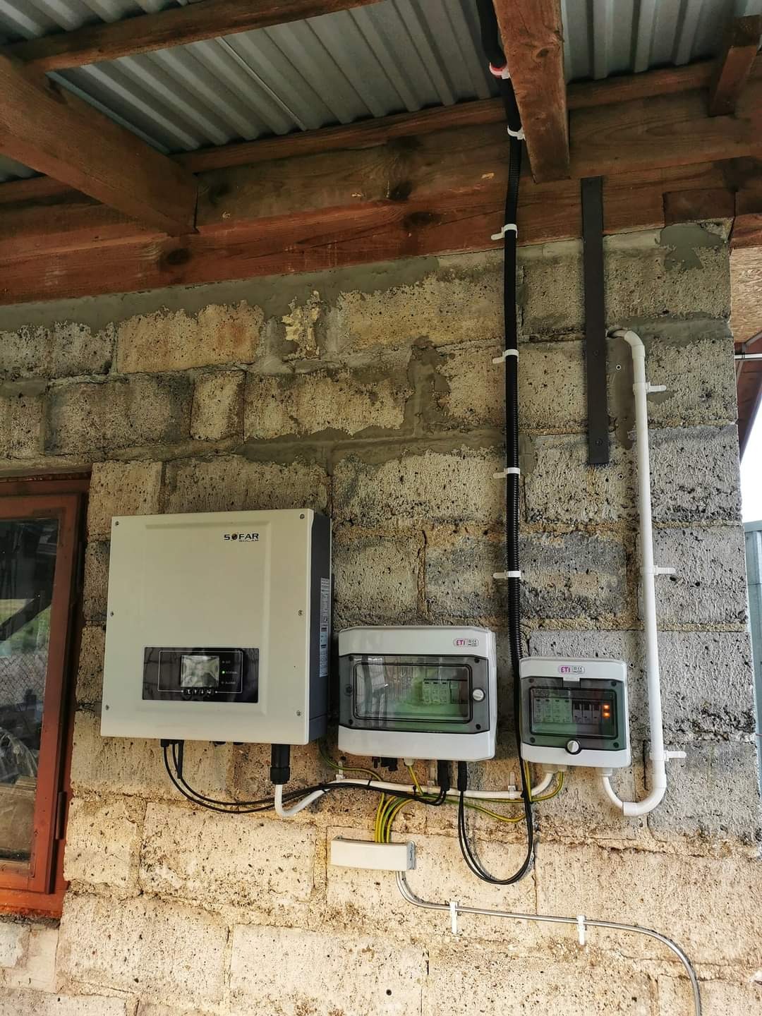 Instalacja paneli fotowoltaicznych: falownik SOFAR i dwie skrzynki zabezpieczające ETI zamontowane na szarej ścianie z bloczków betonowych pod zadaszeniem z blachy falistej.