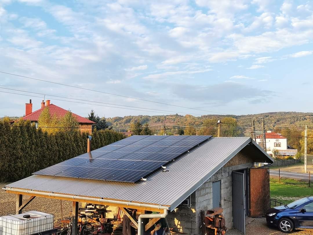 Instalacja paneli słonecznych na dachu garażu z widokiem na wiejski krajobraz i błękitne niebo z chmurami.