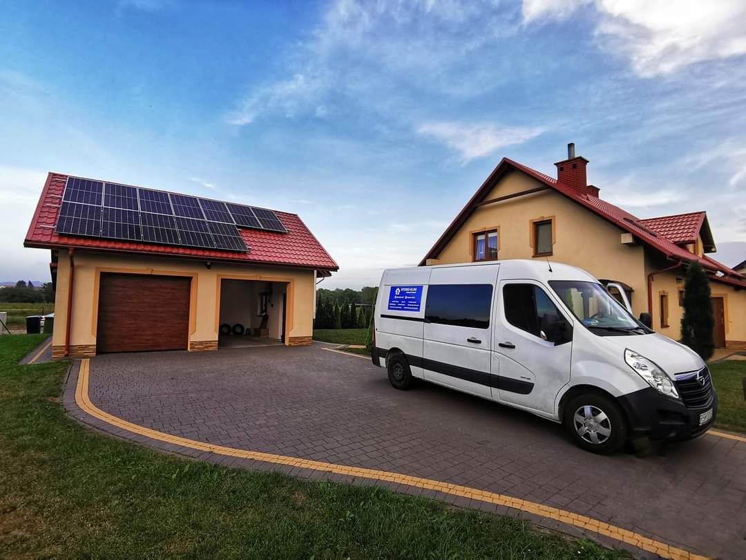 Biały van serwisowy zaparkowany przed garażem z panelami słonecznymi na dachu, w tle dom jednorodzinny z czerwonym dachem.