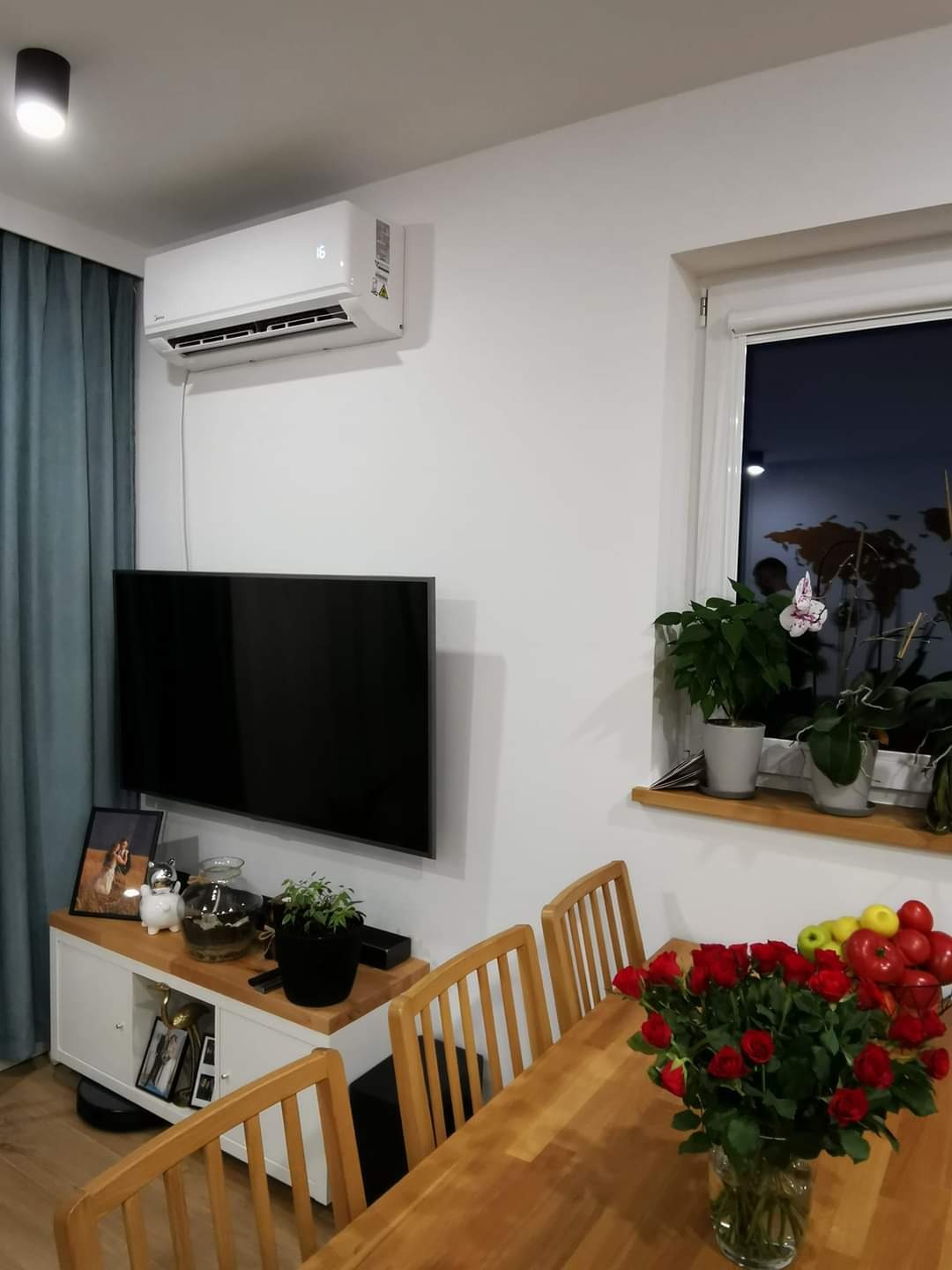 Biały klimatyzator ścienny zamontowany nad telewizorem w jasnym salonie z drewnianym stołem i bukietem czerwonych róż.