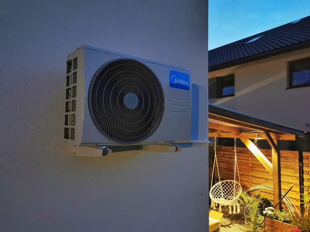 Zewnętrzna jednostka klimatyzacji Midea zamontowana na ścianie domu z widocznym ogrodem i huśtawką w tle, sfotografowana wieczorem przy sztucznym oświetleniu.