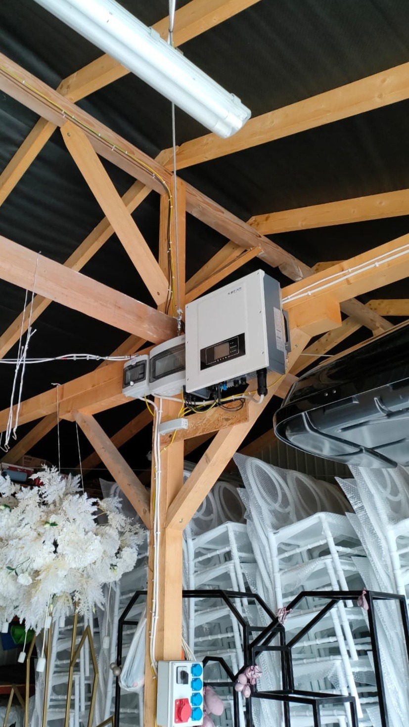 Zamontowany inwerter solarny na drewnianej konstrukcji stropu, widoczne kable, skrzynka elektryczna, lampa i elementy wyposażenia magazynowego w tle.