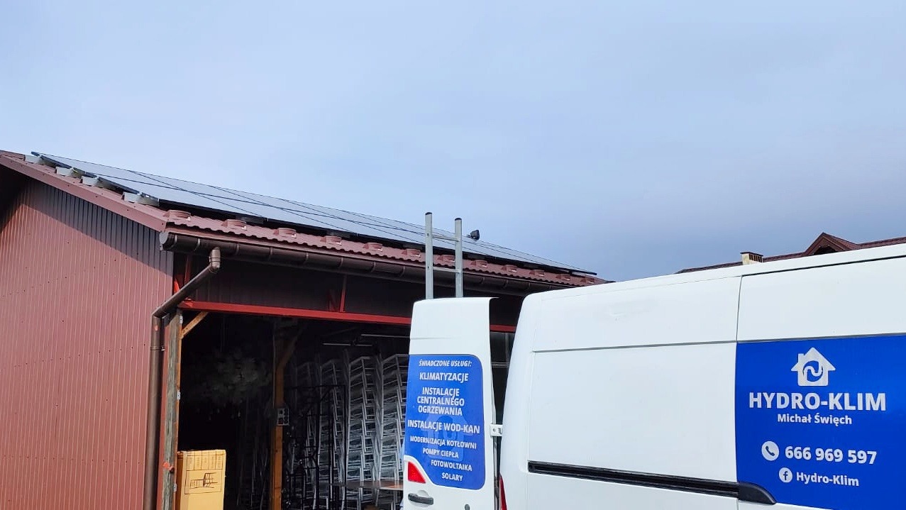 Instalacja paneli fotowoltaicznych na dachu budynku, widoczny samochód firmy Hydro-Klim z reklamą usług klimatyzacji, instalacji centralnego ogrzewania i fotowoltaiki.