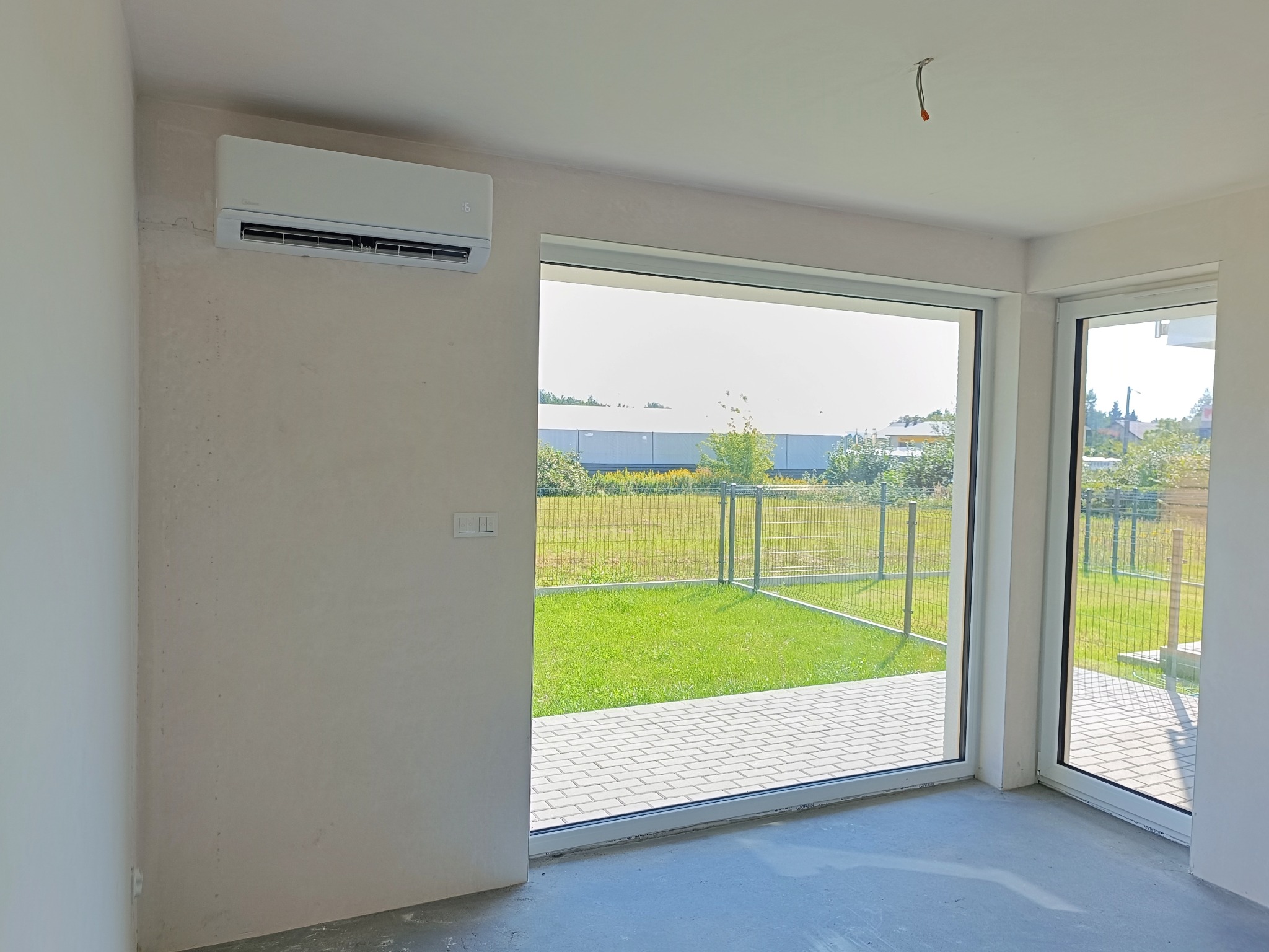 Wnętrze pokoju z zamontowanym klimatyzatorem Midea na ścianie, widok na zewnątrz przez duże okno na trawnik i ogrodzenie, surowy stan wykończenia ścian.