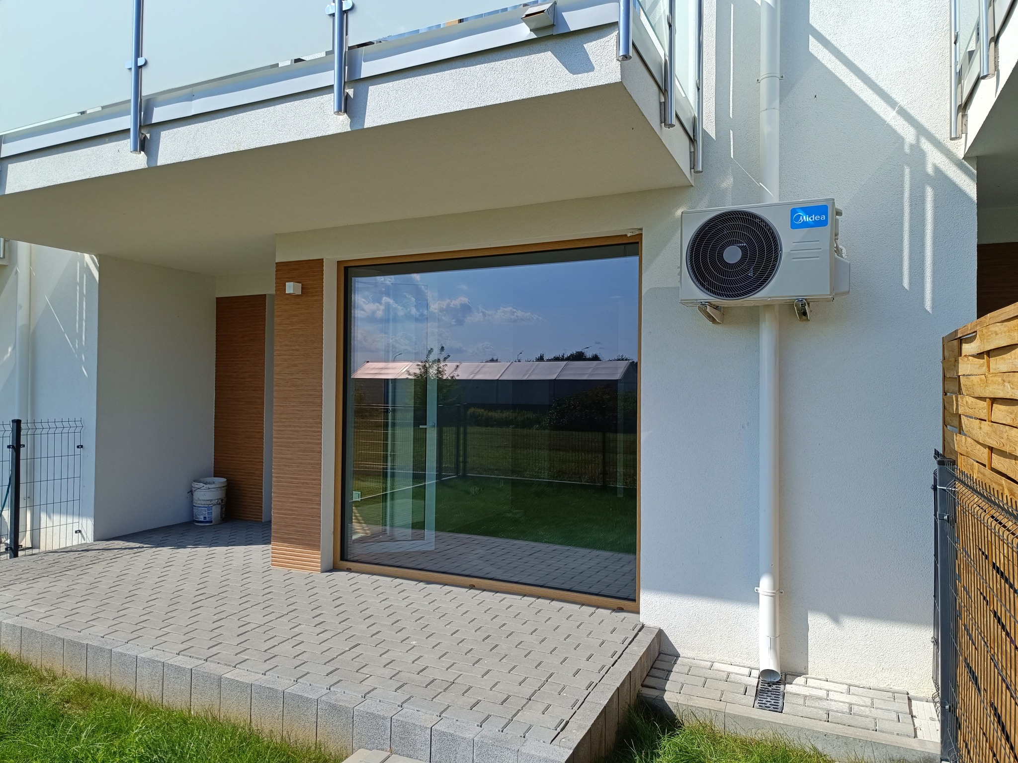 Zewnętrzny klimatyzator Midea zamontowany na białej elewacji domu z dużym oknem i brukowanym tarasem, widoczna rura odprowadzająca skropliny.