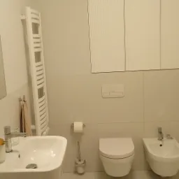 Jasna, minimalistyczna łazienka z białą ceramiką: umywalka na postumencie, toaleta podwieszana i bidet. Na ścianie grzejnik drabinkowy i szafki z ryflowanymi frontami.