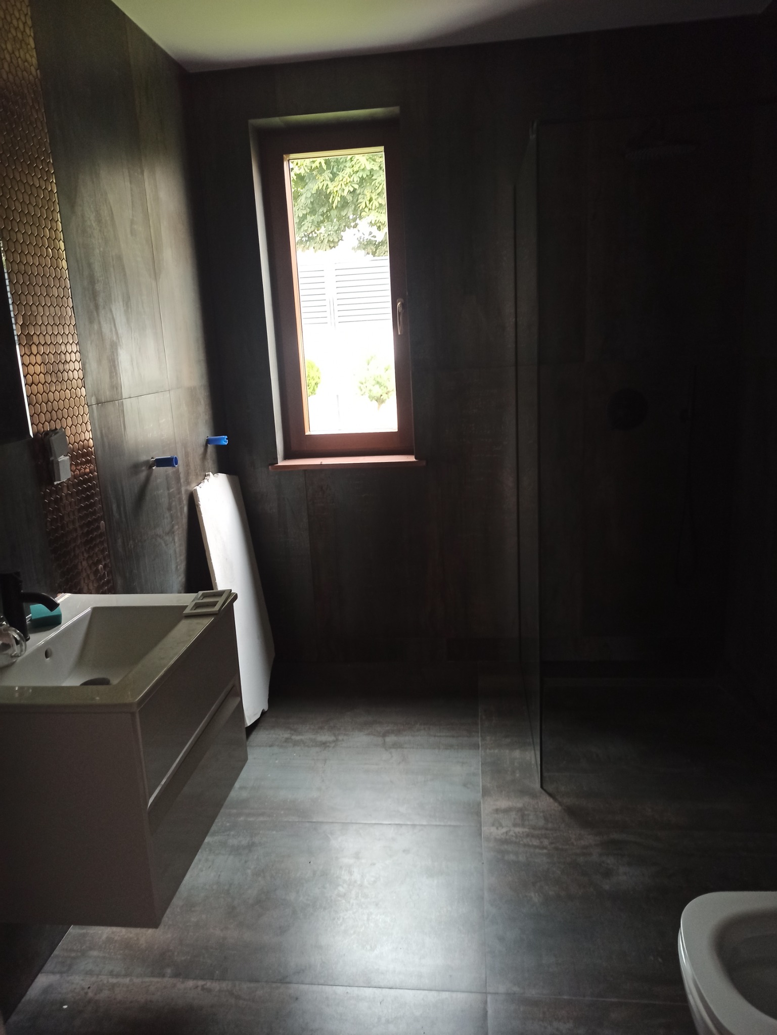 Wnętrze łazienki w trakcie prac wykończeniowych, widoczna umywalka z szafką, fragment kabiny prysznicowej z ciemnymi płytkami, okno z widokiem na zieleń oraz płyta gipsowo-kartonowa oparta o ścianę.